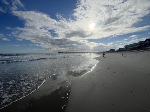 Beach tide recedes reflecting stretching clounnds over and water, pier far in distance, a parent and child silhouetted mid round, along horizon line ,